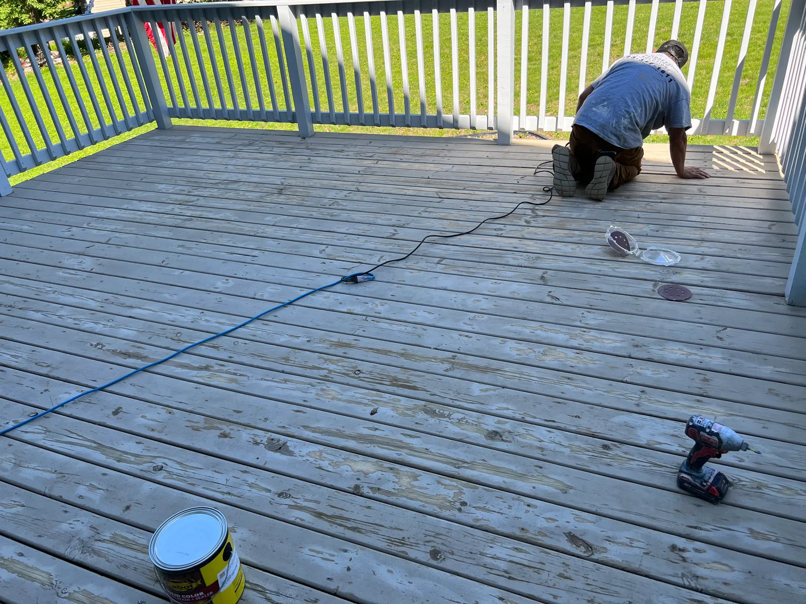 Worker applying gray-blue stain to wooden deck boards with paint can and power drill visible