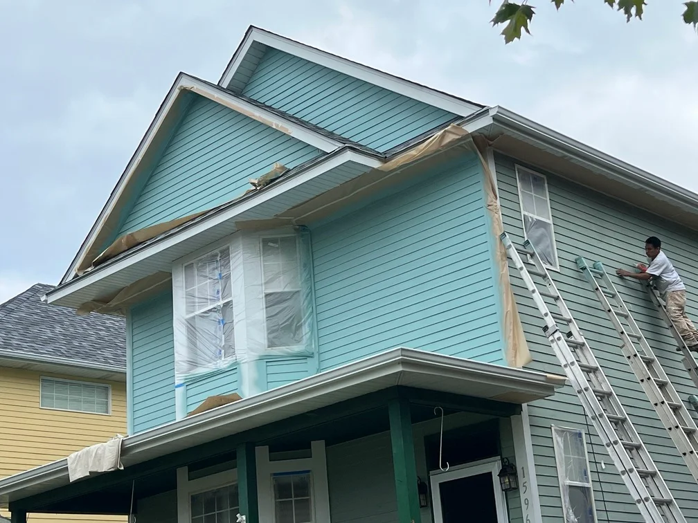 Two-story house with light blue painted siding, protected windows, painter on ladder during exterior painting