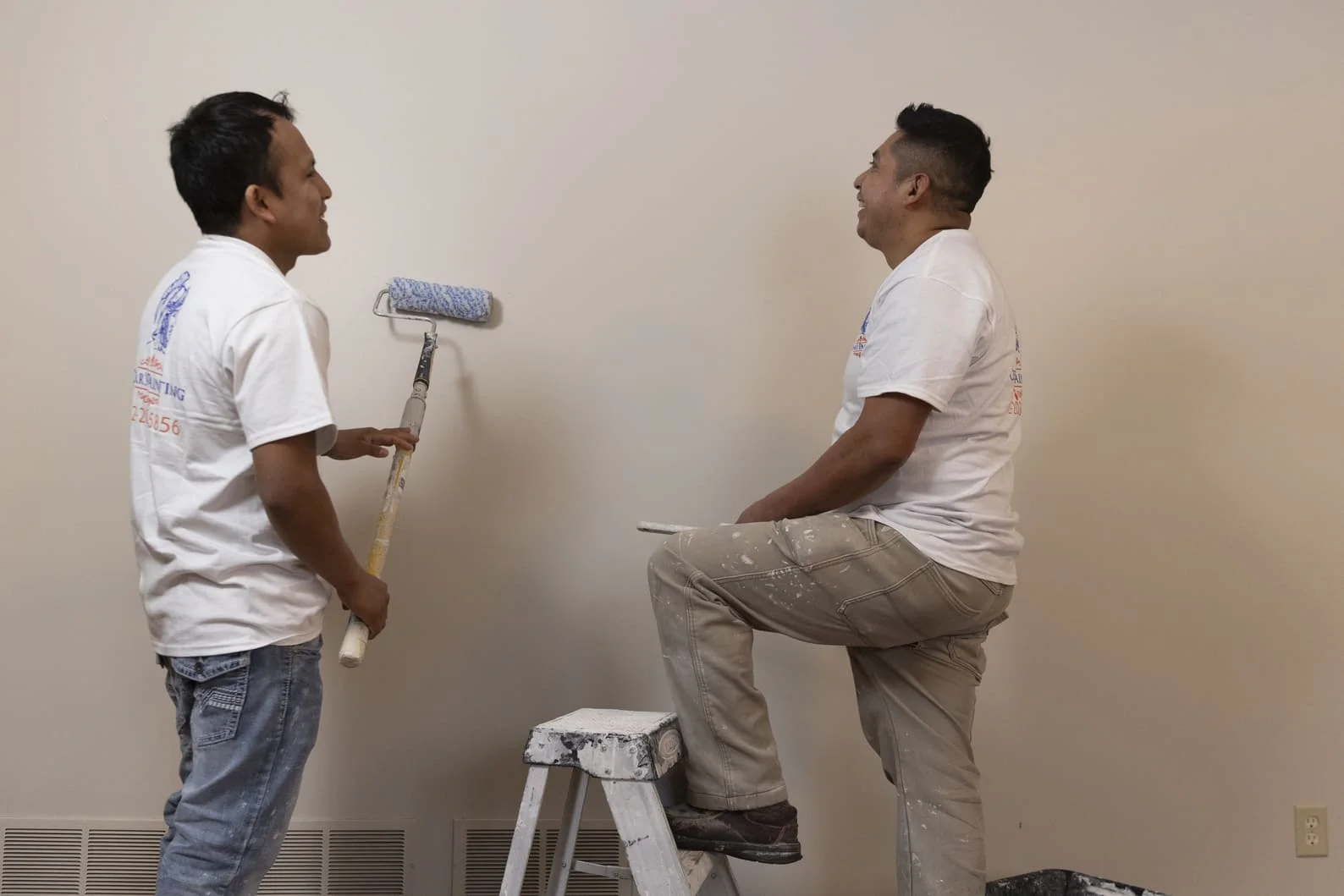 Two painters applying paint to interior wall with roller and brush