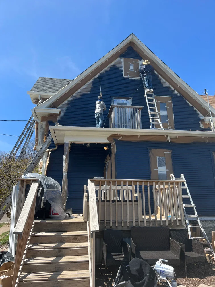 Two-story house with dark blue vinyl siding, painters on ladders working on gable and upper exterior