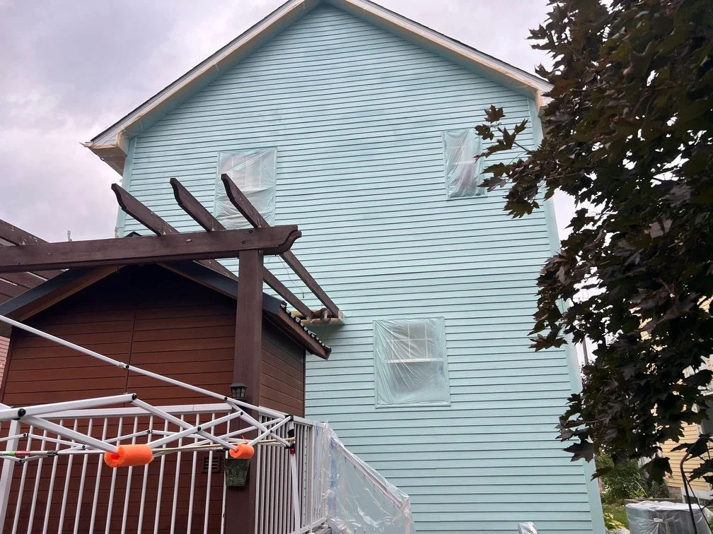 Light blue painted vinyl siding on two-story house with white deck railing and dark brown pergola