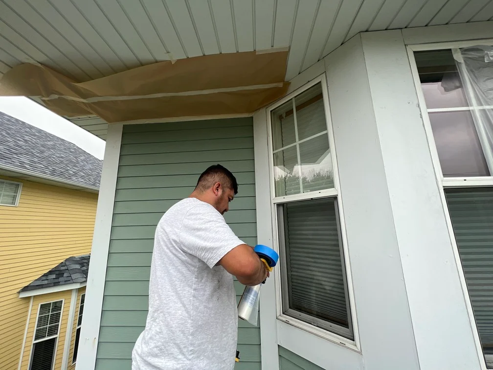 Painter applying sage green exterior paint to horizontal lap siding with white trim and soffit