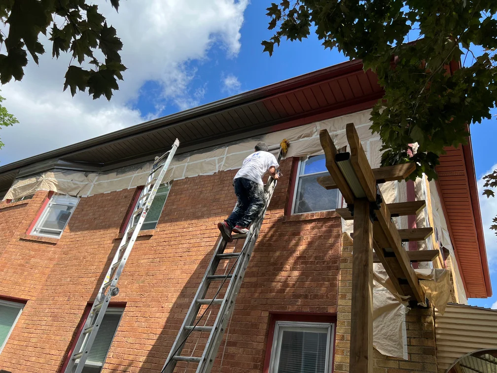 Painter on ladder applying paint to brick exterior wall with protective coverings and masking