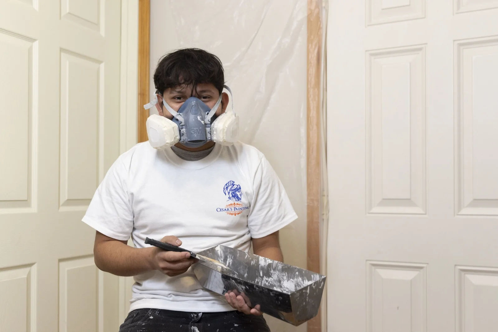 Painter with respirator mask holding putty knife and paint tray during interior wall preparation