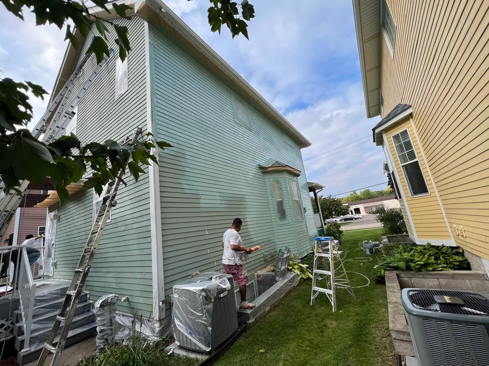 Painter applying exterior paint to light green vinyl siding on residential home with protective coverings