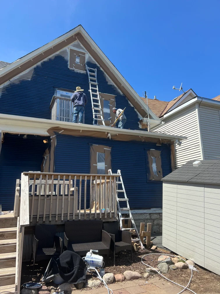 Two-story house with dark blue painted siding, workers on ladders painting upper gable and exterior walls