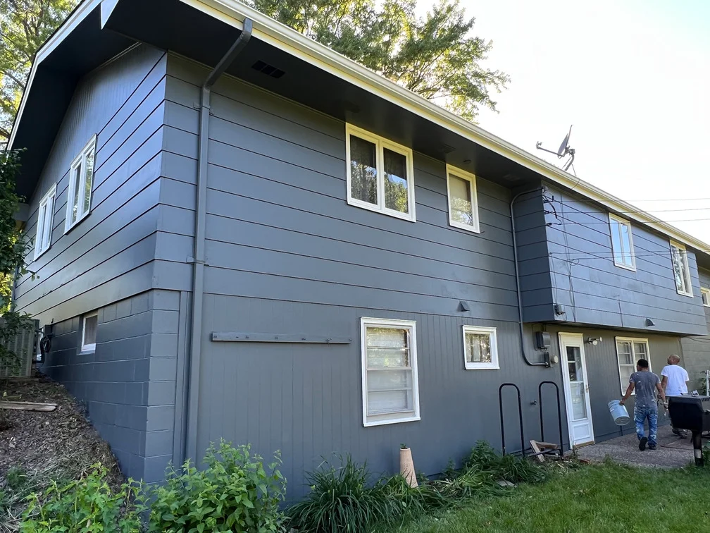 Two-story house with blue-gray horizontal siding, white trim windows, and dark roof overhang