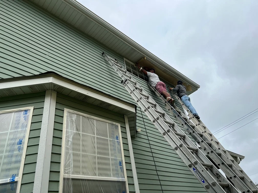 Painter applying finish to green vinyl siding and white soffit trim on residential home exterior