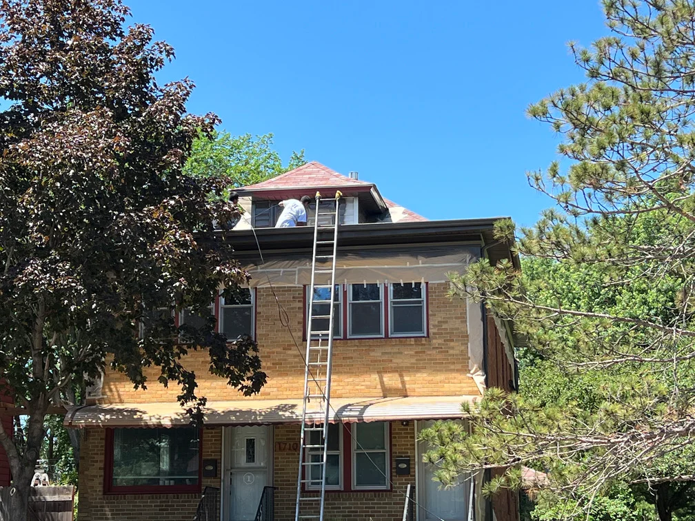 Two-story brick house exterior painting in progress with ladder and protective coverings