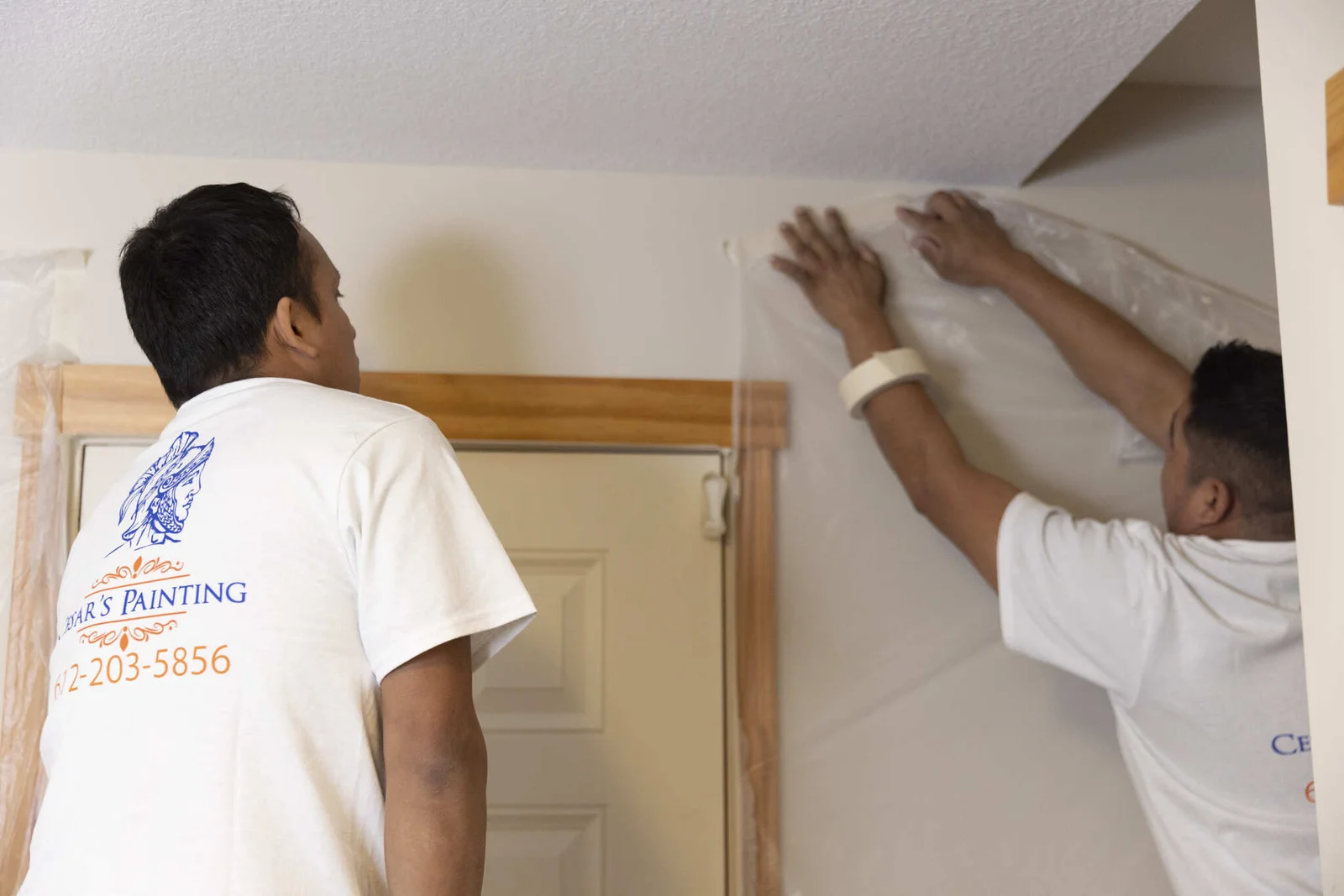 Two painters applying white paint to interior drywall with wooden door frame trim visible