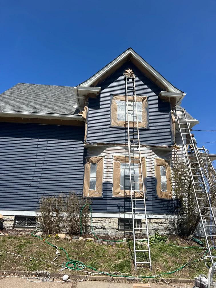 Two-story house with vinyl siding, masked windows, and ladders set up for exterior painting work